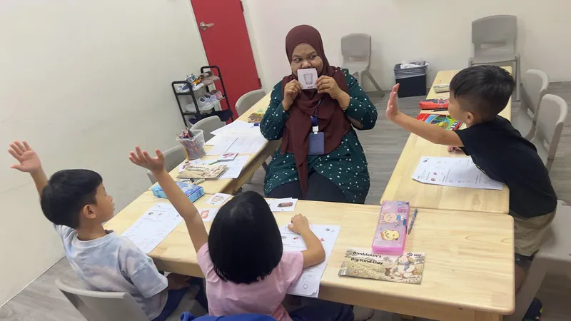 A teacher holding up a flashcard while children around the table raise their hands
